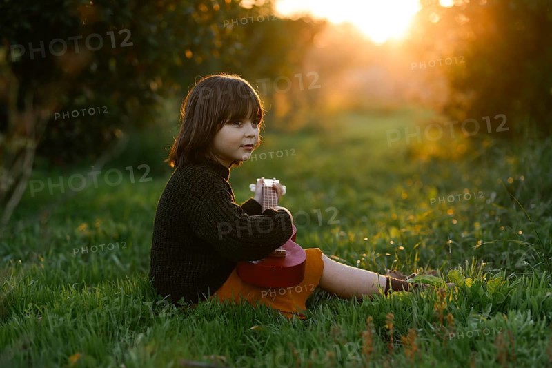 A child sits on lush green grass with orange trees holding a small ukulele, with the warm glow of the setting sun illuminating the tranquil scene, creating a peaceful atmosphere in nature