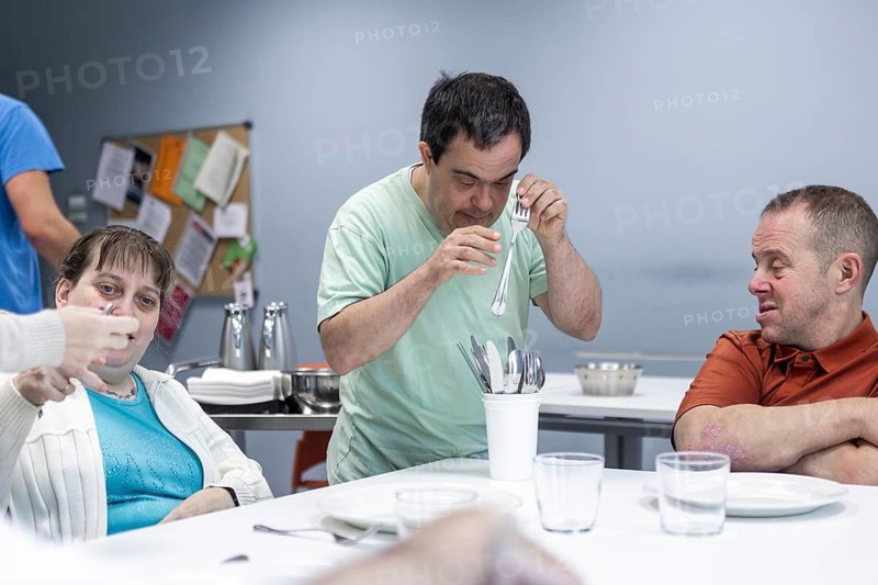 Individuals with intellectual disabilities engage in setting the table, organizing utensils and plates. Their interaction highlights inclusion, independence, teamwork, and life skills development