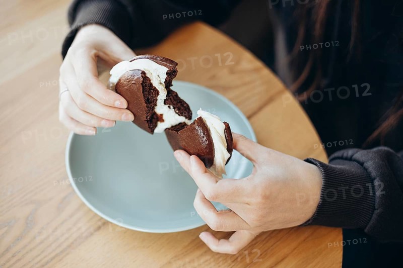 Close-up of hands breaking a chocolate whoopie pie filled with cream, set on a light plate and wooden table. A perfect dessert moment captured in natural lighting