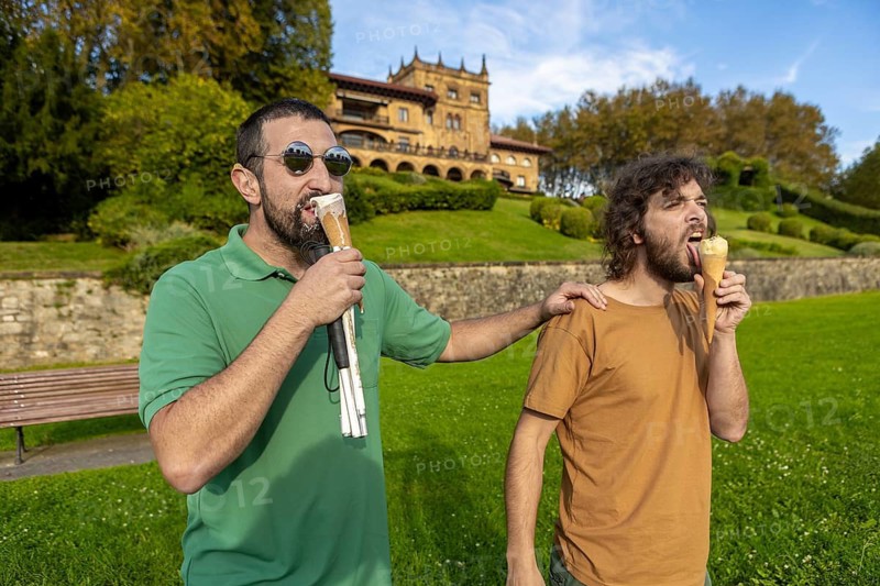 Two friends enjoy ice cream in a sunny park One is blind and uses a cane and sunglasses, symbolizing companionship and trust in a serene outdoor setting