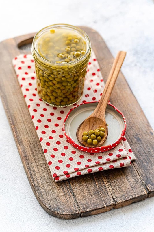 A jar of green homemade organic peas sits on a rustic wooden board, accompanied by a wooden spoon on a polka dot cloth. The scene creates a homely, cozy kitchen vibe