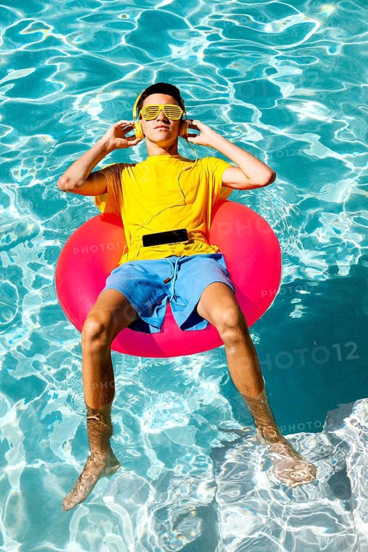 A teenage boy wearing a yellow shirt and sunglasses enjoys music while floating on a pink inflatable in a bright blue swimming pool, epitomizing relaxation and summer vibes