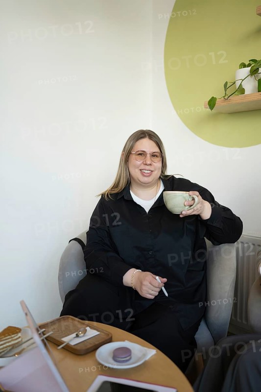 A woman in a black outfit relaxes with a cup of coffee in a bright, stylish cafe. She sits comfortably, surrounded by desserts on the table, creating a welcoming ambiance