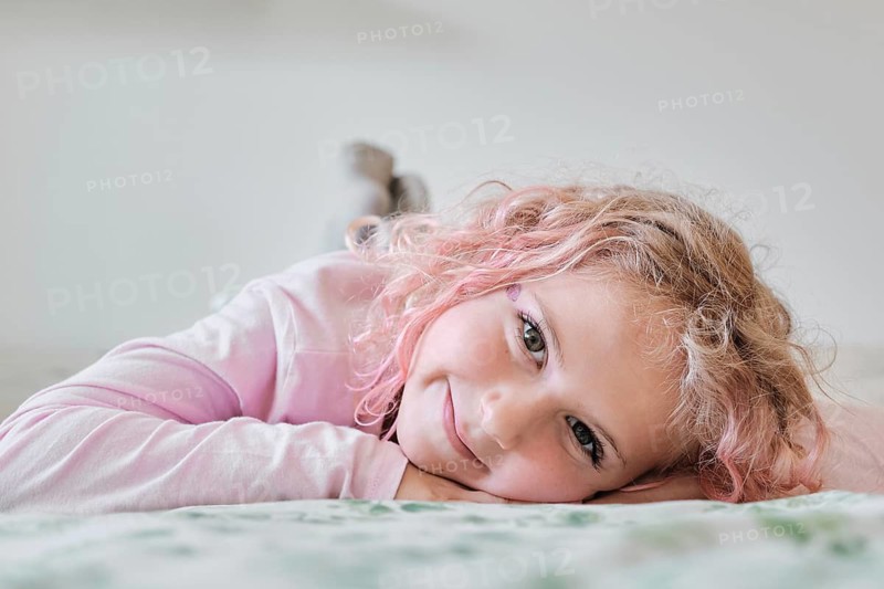A young child with curly hair lies on a bed, smiling gently. The child wears a light sweater, creating a cozy and serene atmosphere at home. The soft focus enhances warmth