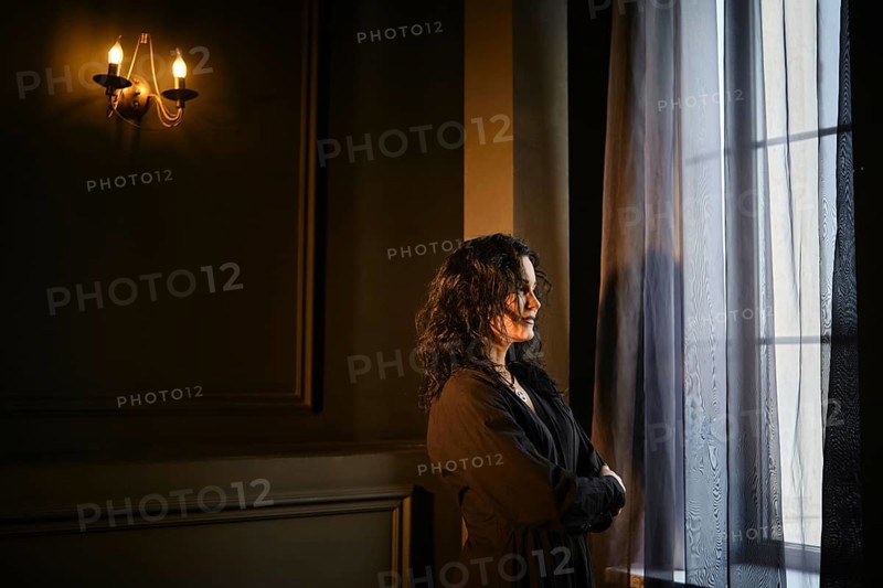 A woman stands by a window in a dark room with soft light from a wall lamp. She gazes outside, lost in thought. The curtain sways slightly, adding a sense of movement to the quiet space