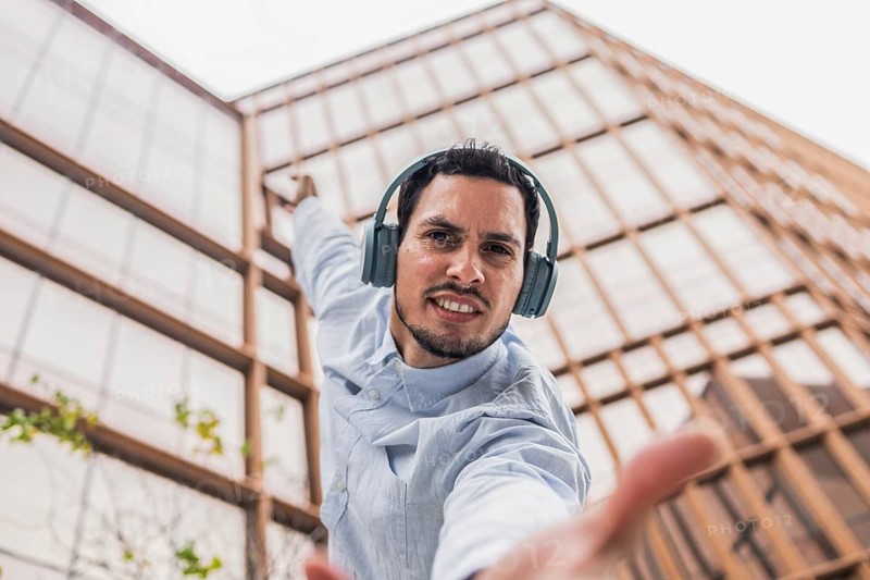 A young Cuban man in a light blue shirt, wearing headphones, strikes a dynamic dance pose. He smiles confidently against a backdrop of modern city architecture