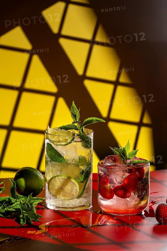 Two refreshing mojito cocktails displayed on a vibrant red surface with a bold yellow background. The classic mojito features lime and mint, while the strawberry mojito is adorned with fresh strawberries