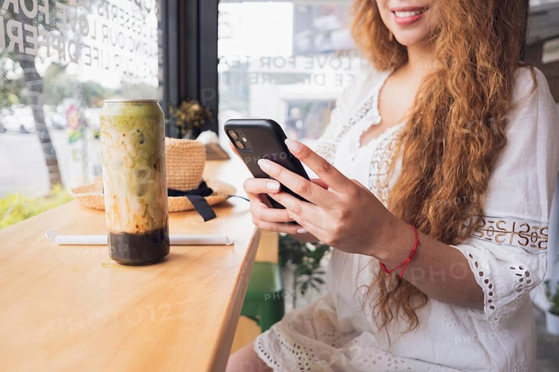 Young American Latin woman enjoying a refreshing drink while using her smartphone in a cozy cafe. The scene captures a modern lifestyle moment with leisure