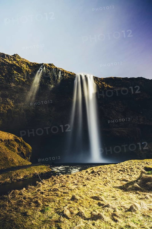 Iconic Seljalandsfoss waterfall in Iceland plunges over mossy cliffs, glowing in golden sunlight under a clear sky. The long exposure captures the ethereal flow of water in motion