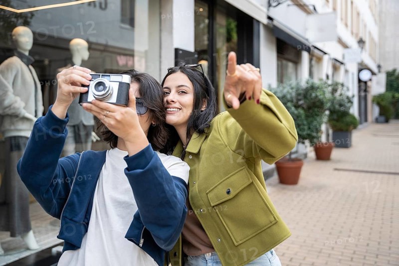 A lesbian couple enjoys a day out, capturing joyful photos with a vintage camera in an urban setting, surrounded by shops and greenery