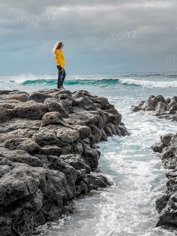 A woman in a yellow jacket stands on rugged volcanic rocks by the ocean, gazing at the waves under a cloudy sky in Timanfaya National Park, Lanzarote, Canary Islands