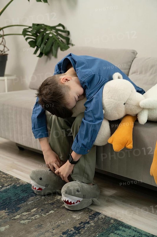 A boy in a blue shirt and green pants sits on a sofa cuddling a plush toy and wearing shark slippers. A relaxed indoor scene with cozy toys and home atmosphere
