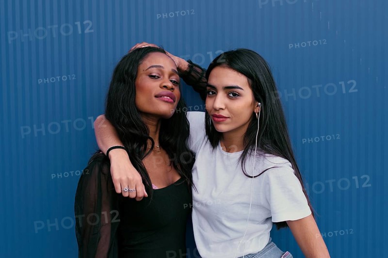 Two young friends pose against a blue background, showcasing friendship and happiness. One wears a white T shirt, the other a black top, both smiling warmly