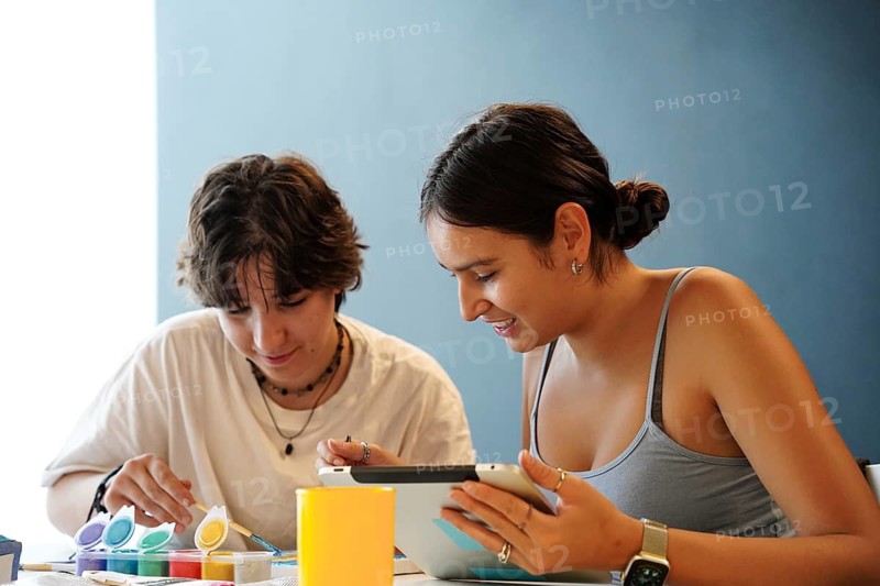Two teenagers, one non-binary and the other female, are focused on painting and using a digital tablet in a classroom setting, symbolizing a blend of traditional and modern educational tools