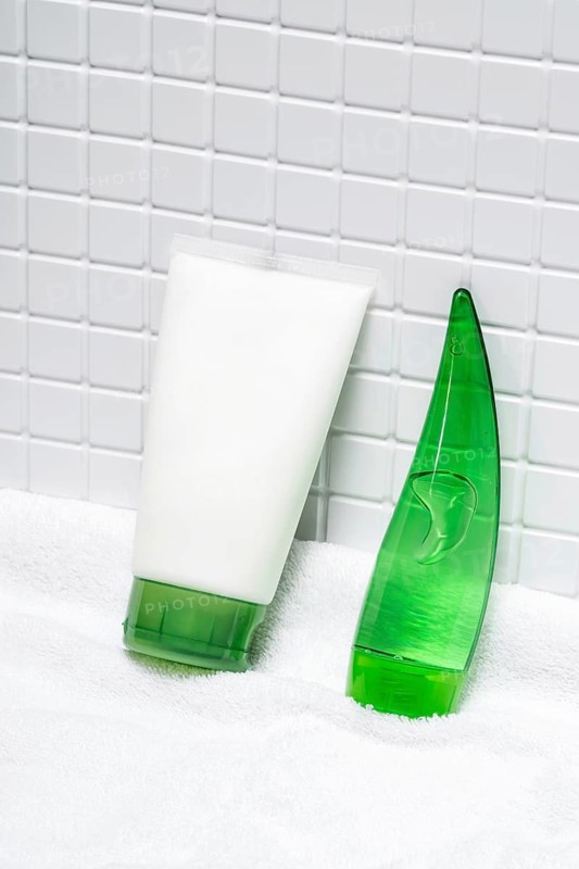 Body care cream and aloe vera gel sit on a fluffy white towel against a white tiled background in a modern, clean bathroom setting