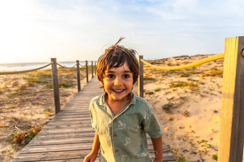 Happy little boy smiling at camera while walking on a wooden boardwalk by sand dunes and the atlantic ocean at golden hour in seignosse, france, carefree summer joy
