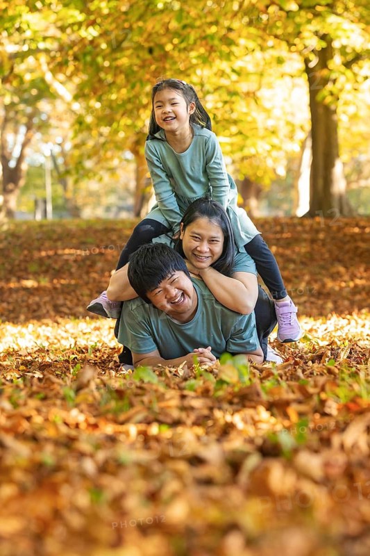 Smiling Asian family enjoying playful moments together outdoors in an autumn park, surrounded by golden leaves. The joy and warmth of family bonds are evident in their expressions