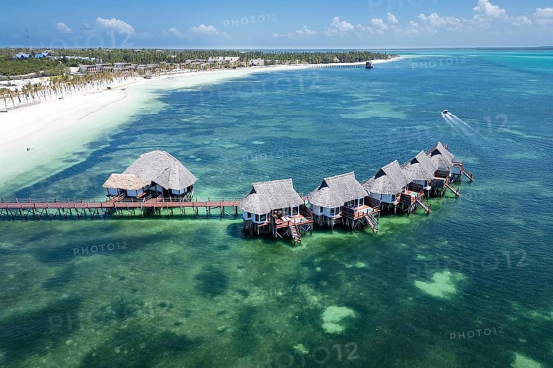 Aerial view of luxury overwater bungalows with thatched roofs and a wooden pier surrounded by emerald ocean and white sand beach in Zanzibar