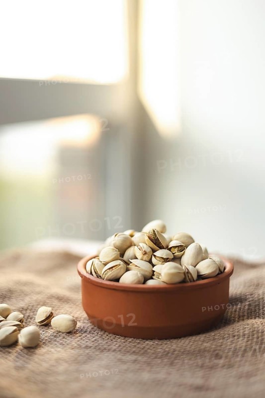 A rustic terracotta bowl filled with pistachios sits on a woven fabric. Sunlight streams in from the window, creating a warm and inviting atmosphere perfect for snacking