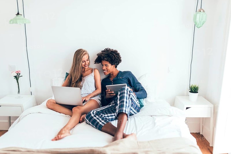 A young couple sits comfortably on a bed in a bright bedroom, engaged with their laptop and tablet. They share a joyful moment, surrounded by a minimalistic, modern decor