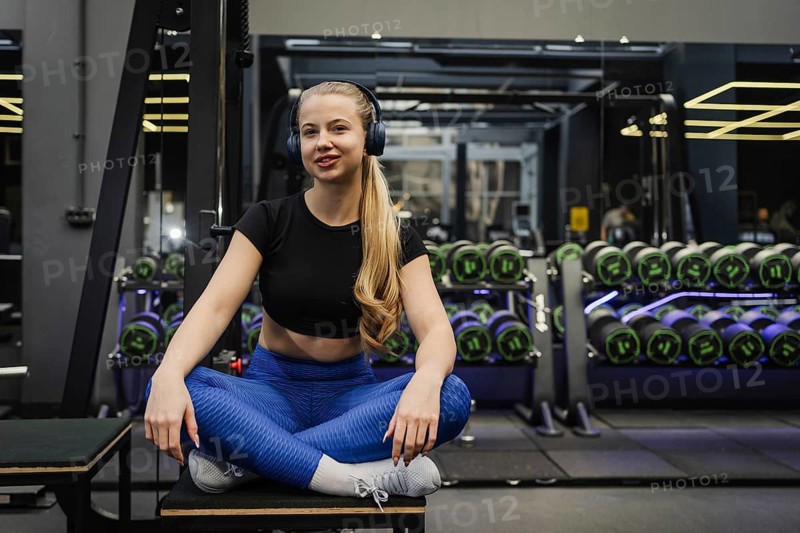 A blonde woman in fitness attire sits on a gym bench, wearing headphones. She appears relaxed after a workout in a modern gym with neatly arranged dumbbells