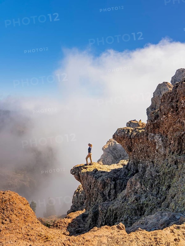 Distant side view of woman standing on the rugged terrain of Pico de las Nieves, Gran Canaria, overlooking a cloud-filled valley
