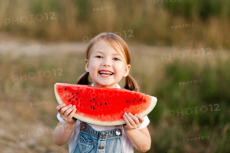 A joyful young child with pigtails holds a big slice of watermelon, smiling brightly during a warm summer day. The outdoor setting enhances the cheerful summer vibe