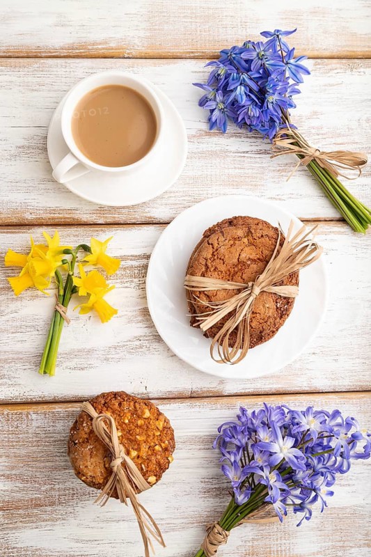 Oatmeal cookies with spring snowdrop flowers bluebells, narcissus and cup of coffee on white wooden background. top view, flat lay, close up, still life. Breakfast, morning, spring concept
