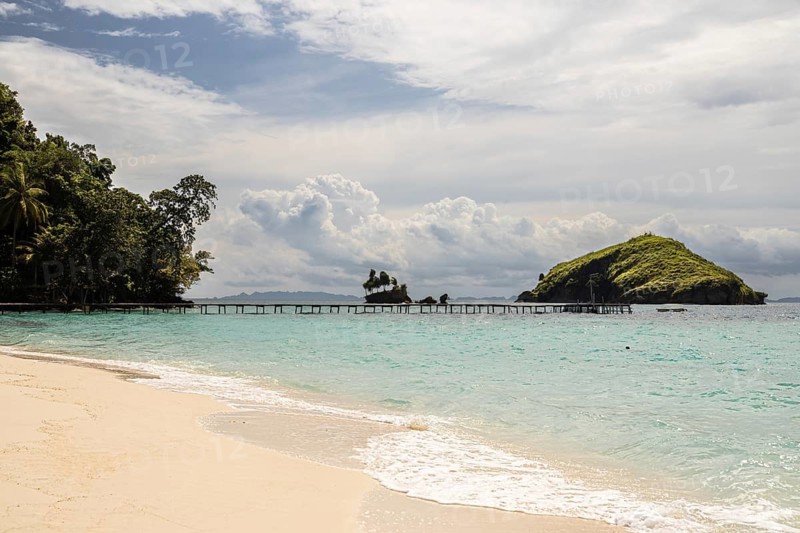 From above, this aerial image captures the serene beach and crystal clear waters of the Raja Ampat Archipelago in Indonesia, featuring a verdant island and a wooden pier stretching into the turquoise sea