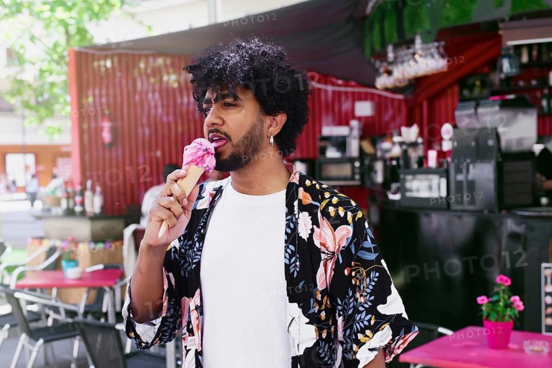 A man enjoys a refreshing ice cream cone on a warm summer day, standing in a vibrant outdoor setting. The bright sun highlights his relaxed demeanor amid a lively backdrop