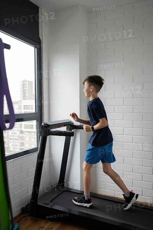 A young boy in a blue sports outfit is jogging on a treadmill inside a bright room with white brick walls and a large window, staying active and healthy indoors