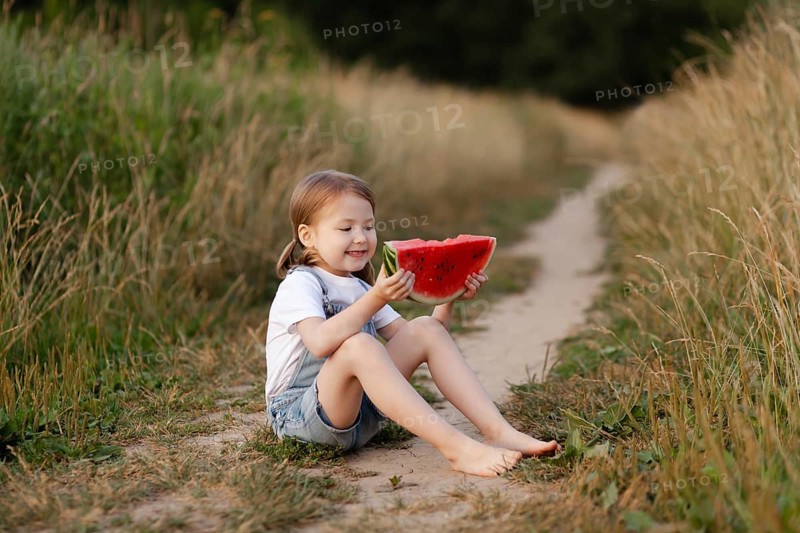 A joyful child sits outdoors, savoring a fresh watermelon slice on a sunny summer day. The scene captures the essence of childhood and summertime fun in a natural setting