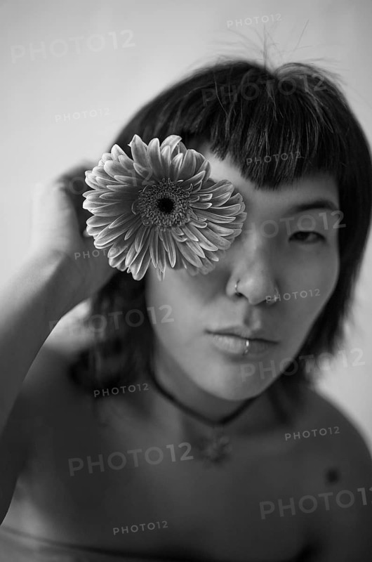 Black and white portrait of an Asian woman holding a flower to her eye, highlighting her serene and artistic expression. The image conveys a sense of calm and creativity