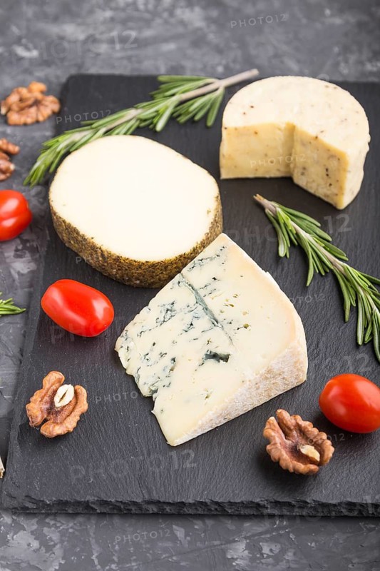 Blue cheese and various types of cheese with rosemary and tomatoes on black slate board on a black concrete background. Side view, close up, selective focus