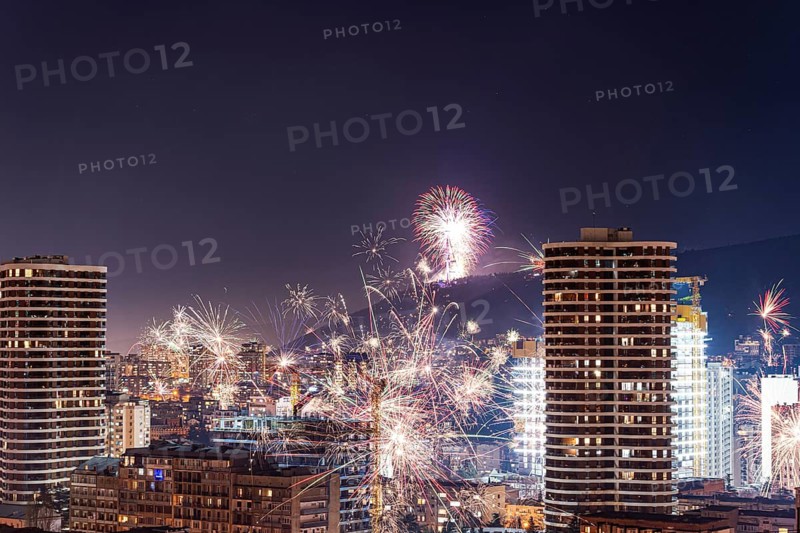 Vibrant fireworks light up the night sky over Tbilisi's downtown, illuminating the cityscape with colorful bursts. Skyscrapers and urban lights offer a stunning backdrop