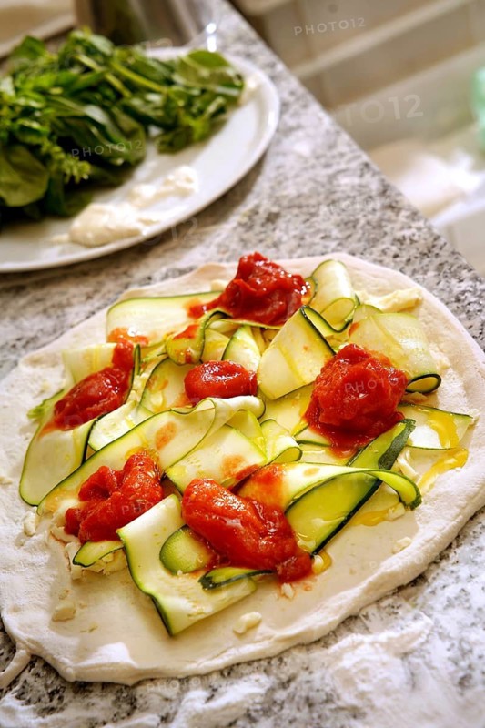 Cropped unrecognizable view of fresh pizza preparation with slices of zucchini and dollops of tomato sauce on raw dough, set on a marble countertop