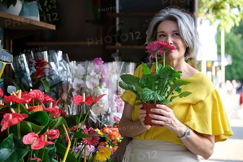 A mature woman with gray hair enjoys the scent of pink flowers at an outdoor flower market, radiating joy and appreciation for nature's beauty, while wearing a yellow blouse