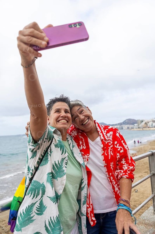 A happy couple of women taking a selfie with a smartphone on a cloudy beach day, dressed in summer attire, capturing precious holiday moments
