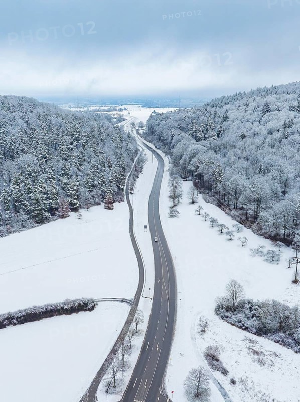 Snow-covered road through a wintry landscape with wooded hills and few moving cars, Aidlingen, Böblingen district, Germany