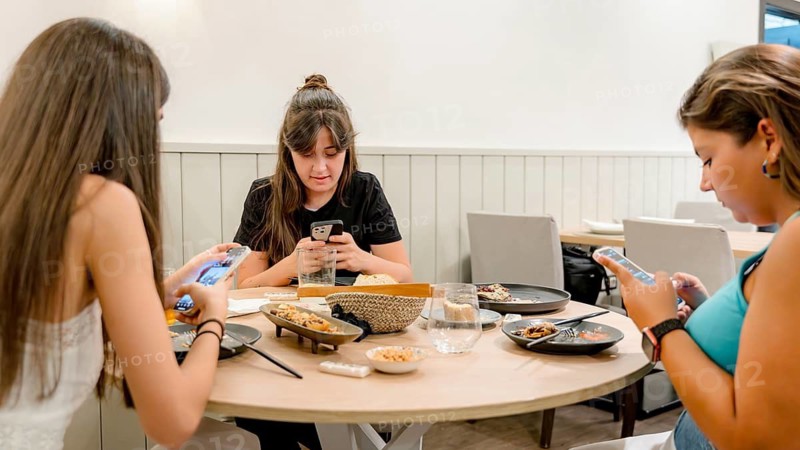 Three young women are engrossed in their smartphones while sitting at a stylish restaurant table. The table is set with a variety of dishes, creating a casual dining atmosphere
