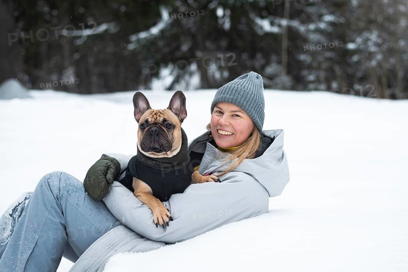 A cheerful person enjoys a winter day playing in the snow with a cute dog. Dressed warmly, they sit in a snowy field, capturing the essence of outdoor fun and companionship