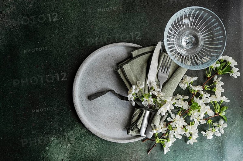 Aerial view of an elegant table setting with white blossoms, a glass, and cutlery on a ceramic plate, all laid on a muted green background for a minimalist look