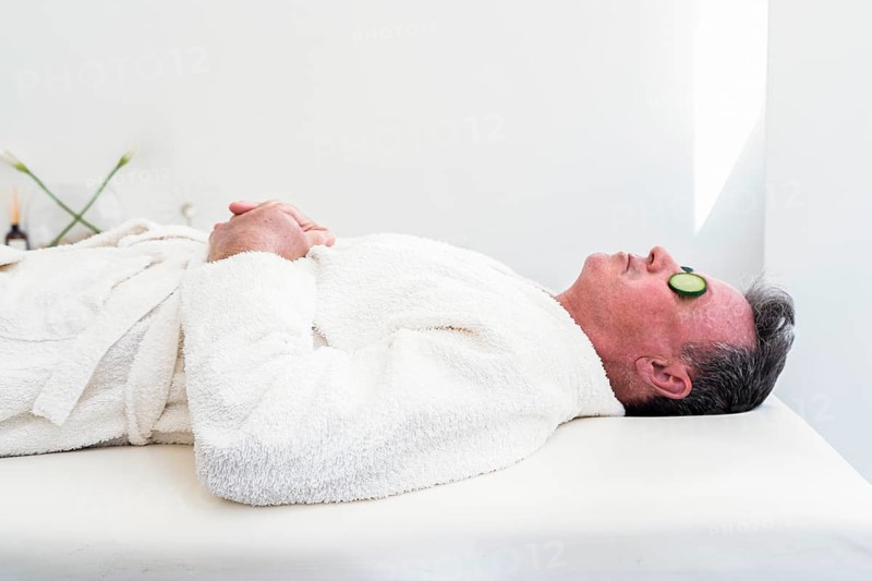 A mid-adult man enjoys a calming spa session. He lies down, eyes covered with cucumber slices, wearing a cozy white robe on a plush spa bed