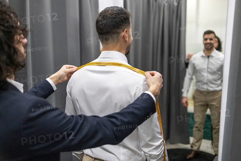 A professional tailor measures a client shoulder width using a tape measure in a fitting room. Both are well dressed, highlighting the tailoring process and personalized service
