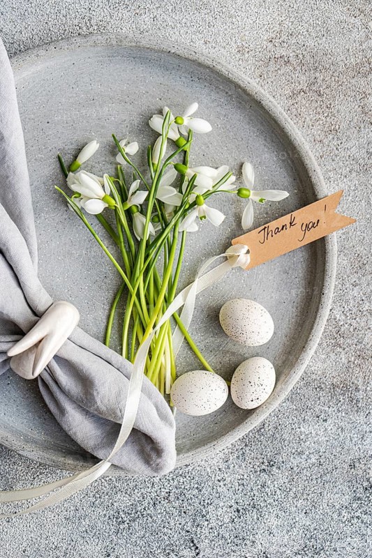 Top view of fresh snowdrops and speckled eggs arranged in a concrete dish, accompanied by a Thank you! tag, symbolizing gratitude and the arrival of spring