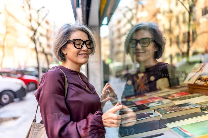Mature woman with gray hair and eyeglasses smiling while carefully looking at a book display in a shop window, enjoying her leisure time on a city street