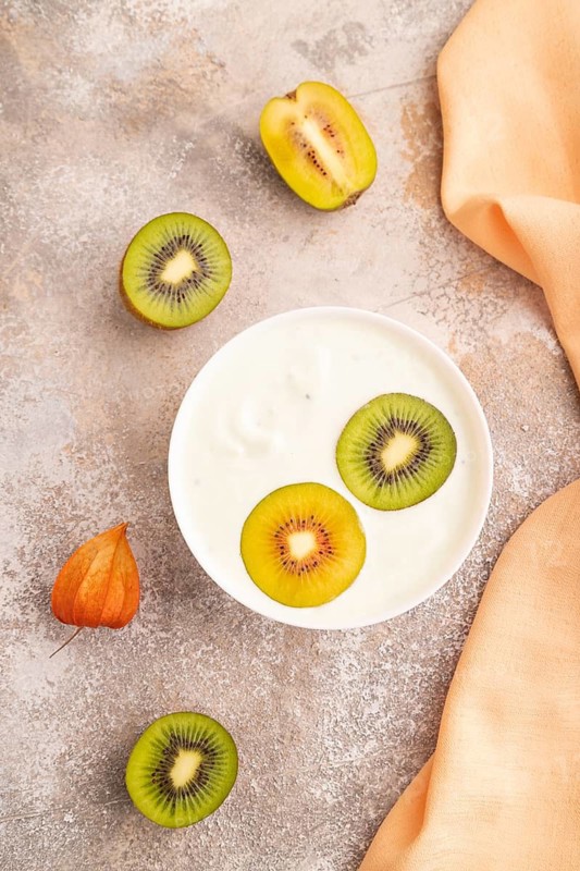 Yogurt with kiwi in white bowl on brown concrete background and orange linen textile, top view, flat lay, close up, minimalism