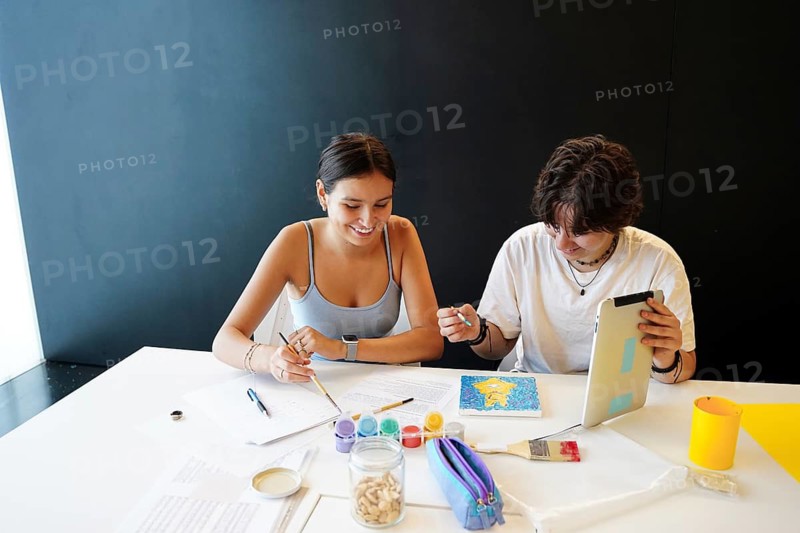 Two teenagers, one non-binary and one female, participate in artistic activities at school. They are focused on painting and using various study materials, including a digital tablet, enhancing their creativity and teamwork