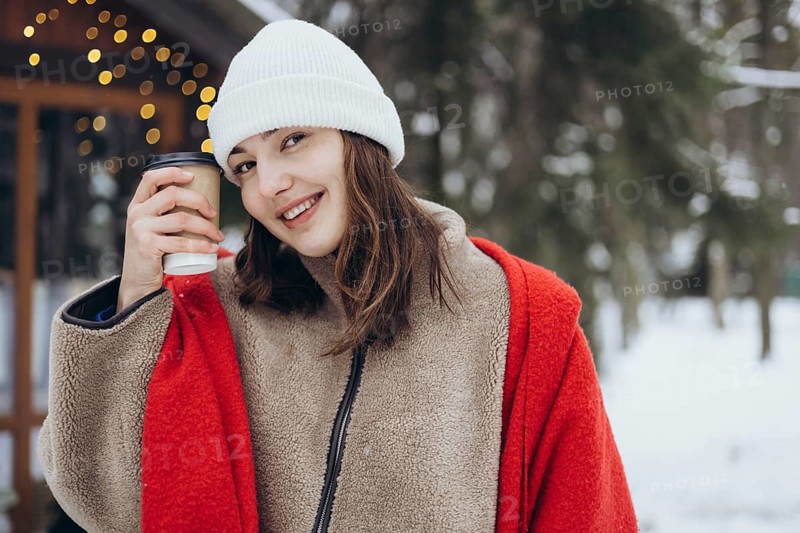 A young woman smiles holding a coffee cup, dressed warmly in a red scarf and white beanie against a snowy background with soft focus lights