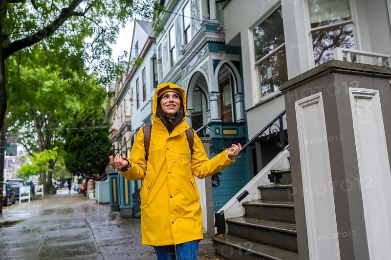 Woman in a vibrant yellow raincoat and backpack standing on a city sidewalk in san francisco, smiling and looking up, with victorian houses lining the wet street on a cloudy day
